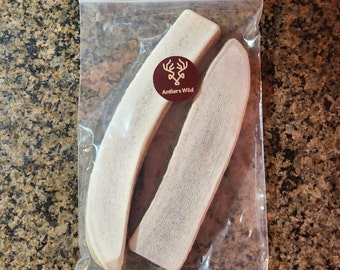 May include: Two light-colored antler chews in a clear plastic bag. A round sticker with the text "Antlers Wild" and a deer head logo is affixed to the bag. The chews have a curved shape and a rough texture.
