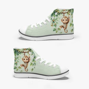 May include: Two high-top canvas sneakers. The sneakers are light green with white laces and soles. Each shoe features a watercolor illustration of a monkey hanging from a tree branch with green leaves and small orange fruits. The design is repeated on both shoes.