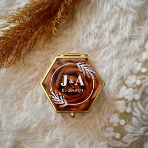 May include: A hexagonal ring box with a clear lid and a gold metal frame. The box contains a faux rose with a white floral design and the text "J&A 05-20-2023".