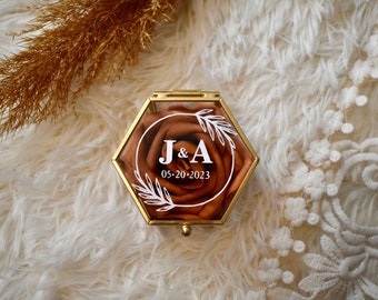 May include: A hexagonal ring box with a clear lid and a gold metal frame. The box contains a faux rose with a white floral design and the text "J&A 05-20-2023".