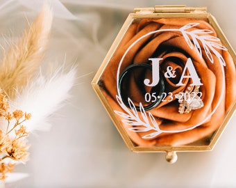 May include: A gold hexagonal ring box with a clear lid, containing two wedding bands and a diamond engagement ring. The box is lined with a peach-colored fabric rose and has the initials "J&A" and the date "05.23.2022" printed on it.