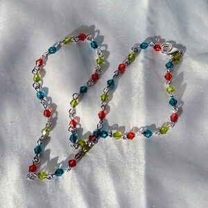 Puede incluir: Un delicado collar con una cadena plateada adornada con cuentas de colores en tonos rojos, verdes y azules. El collar tiene un cierre de mosquetón. Las cuentas están espaciadas uniformemente a lo largo de la cadena.