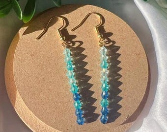 May include: A pair of dangle earrings with gold-colored hooks. Each earring features a strand of small, gradient blue beads, transitioning from light to dark. The earrings are displayed on a round, tan surface, with green foliage in the background.