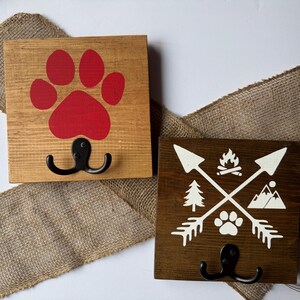 May include: Two square wooden wall hooks featuring black metal double hooks. The left hook has a light brown finish with a red paw print. The right hook has a dark brown finish with a white graphic of crossed arrows, a campfire, a tree, a mountain, and a paw print.