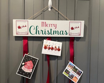 May include: A white wooden Christmas sign with the words "Merry Christmas" in green and red. The sign has two postage stamp designs with Santa and his sleigh. Red ribbons hang below, holding Christmas cards.