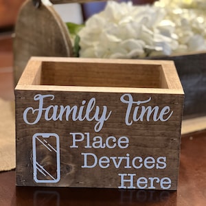 May include: A rectangular brown wooden box with white text that reads Family time Place Devices Here and a white outline graphic of a smartphone. The box has a visible wood grain texture and a hollow interior, designed for holding mobile devices.