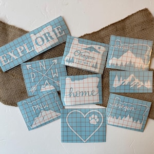 Peut inclure: Plusieurs autocollants en vinyle bleu clair avec des motifs et du texte blancs. Les autocollants présentent des mots comme "EXPLORE" et "HOME", ainsi que des graphiques de montagnes et de boussoles. Les autocollants sont disposés sur une surface en toile de jute.