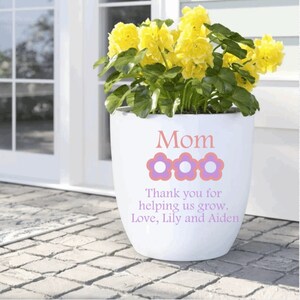 May include: White ceramic planter with the words "Mom, Thank you for helping us grow. Love, Lily and Aiden" in pink lettering. Three pink flowers with purple centers are above the text. A yellow flowering plant is in the planter.