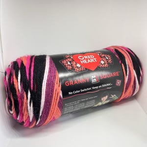 May include: A skein of Red Heart All in One Granny Square yarn featuring a variegated color palette of black, white, bright pink, and orange. The black paper label displays the brand logo, product name, and text including No Color Switchin'.