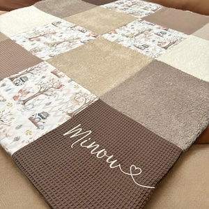 Könnte beinhalten: Eine braune und beige Patchwork-Babydecke mit einem weißen gestickten Namen "Minou" und einem Herz. Die Decke besteht aus verschiedenen Texturen, darunter Waffelpiqué und Plüsch.