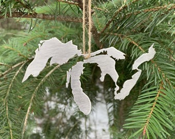 May include: A silver-colored metal ornament shaped like the Great Lakes region, hanging from a natural twine string. The ornament is suspended against a backdrop of green pine needles and a blurred winter scene.