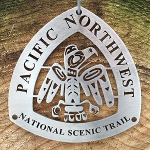 May include: A silver metal ornament shaped like a triangle with a stylized eagle design and the text "PACIFIC NORTHWEST NATIONAL SCENIC TRAIL".