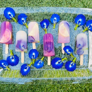 May include: A tray of homemade popsicles with purple and blue hues, decorated with blue butterfly pea flowers. The popsicles are on a clear plastic tray with a textured surface.