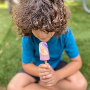 May include: A young person with curly brown hair sits on the grass and holds a purple and pink popsicle with a lemon slice in it.