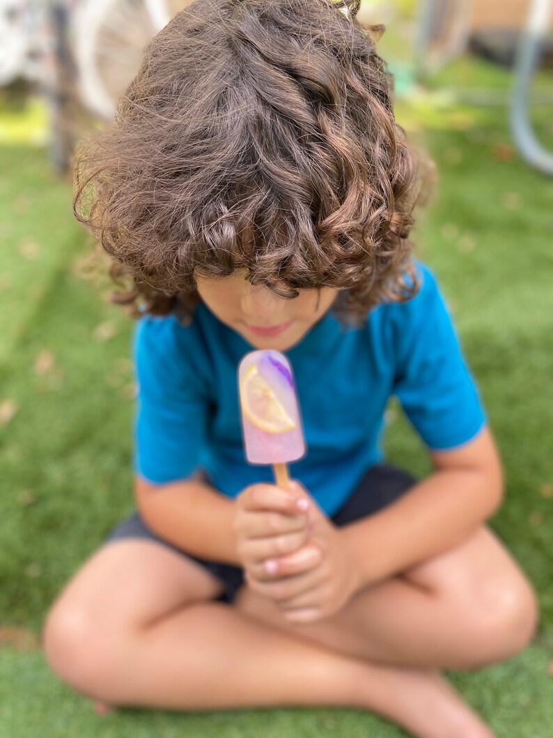 May include: A young person with curly brown hair sits on the grass and holds a purple and pink popsicle with a lemon slice in it.