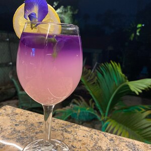 May include: A glass of purple and pink lemonade with a lemon wedge and a blue flower garnish. The drink is in a clear glass with a stem and a wide base.