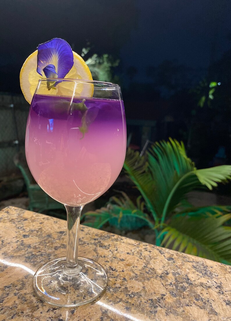 May include: A glass of purple and pink lemonade with a lemon wedge and a blue flower garnish. The drink is in a clear glass with a stem and a wide base.
