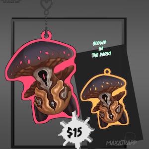 Peut inclure: Un charm en forme de champignon avec un chapeau bleu foncé et une tige marron, souligné en rose. Le charm a un dessus en forme de cœur pour l'accrocher. Le texte "GLOWS IN THE DARK!" est visible. Un accessoire unique.