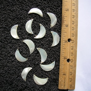 May include: A collection of 12 white mother of pearl half-moon shaped embellishments with gold-toned metal loops. The embellishments are arranged on a black surface next to a wooden ruler.