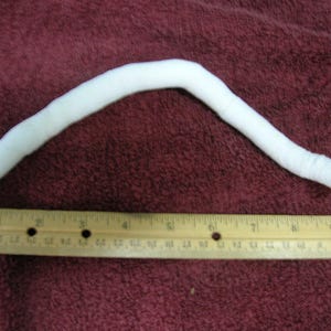 May include: A white, flexible, tube-like object, approximately 25 cm long, is lying on a burgundy fabric surface. A wooden ruler is placed below the object for scale.