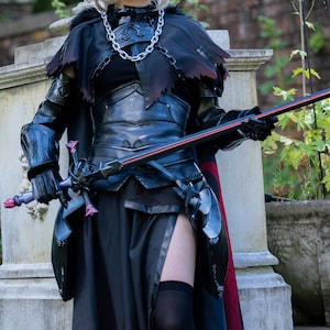 Puede incluir: Una persona con un disfraz de cosplay negro y rojo, sosteniendo una espada. El disfraz incluye un corpiño, una falda y una capa negros. La persona está de pie sobre escalones de piedra en un jardín, con flores y una maceta de piedra en el fondo.