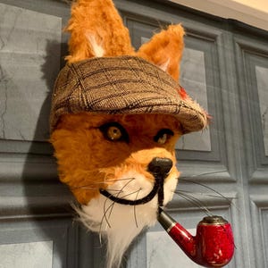 May include: A plush fox head wearing a brown and white checked newsboy cap and holding a red and black pipe. The fox has a white beard and moustache.
