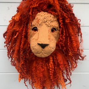 Puede incluir: Una cabeza de león de punto con una melena roja y esponjosa. La cara del león es beige con ojos marrones y una nariz negra.