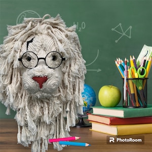 May include: A white yarn lion head with a black lightning bolt scar on its forehead, wearing round black glasses. The lion head is sitting on a wooden table with a globe, a green apple, and a stack of books in the background.