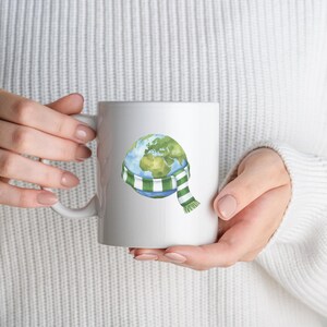 May include: White ceramic mug featuring a watercolor illustration of Earth wearing a green and white striped scarf. The mug is held by a person wearing a white sweater. The Earth illustration shows the continents in green and the oceans in blue.