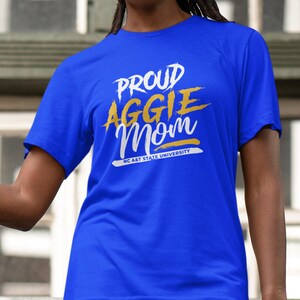 May include: Royal blue t-shirt with the text "PROUD AGGIE Mom" in white and gold lettering. The text "NC A&T STATE UNIVERSITY" is printed below the main text in white. The shirt is a short-sleeved crew neck.