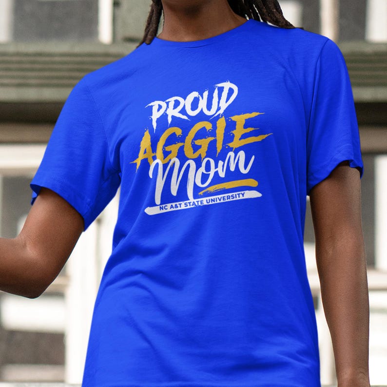 May include: Royal blue t-shirt with the text "PROUD AGGIE Mom" in white and gold lettering. The text "NC A&T STATE UNIVERSITY" is printed below the main text in white. The shirt is a short-sleeved crew neck.