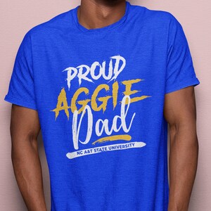 May include: Royal blue t-shirt with the words "PROUD AGGIE Dad" in white and yellow lettering. The text "NC A&T STATE UNIVERSITY" is printed in white at the bottom. The shirt is short-sleeved.