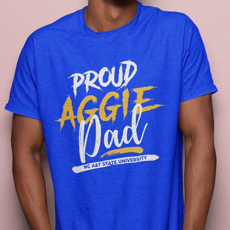 May include: Royal blue t-shirt with the words "PROUD AGGIE Dad" in white and yellow lettering. The text "NC A&T STATE UNIVERSITY" is printed in white at the bottom. The shirt is short-sleeved.
