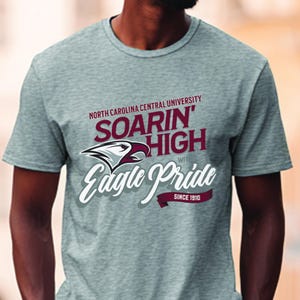 Peut inclure: T-shirt gris avec le texte "NORTH CAROLINA CENTRAL UNIVERSITY" au-dessus de "SOARIN' HIGH" et "Eagle Pride" avec un graphisme d'aigle. Le t-shirt porte &eacute;galement l'inscription "SINCE 1910".
