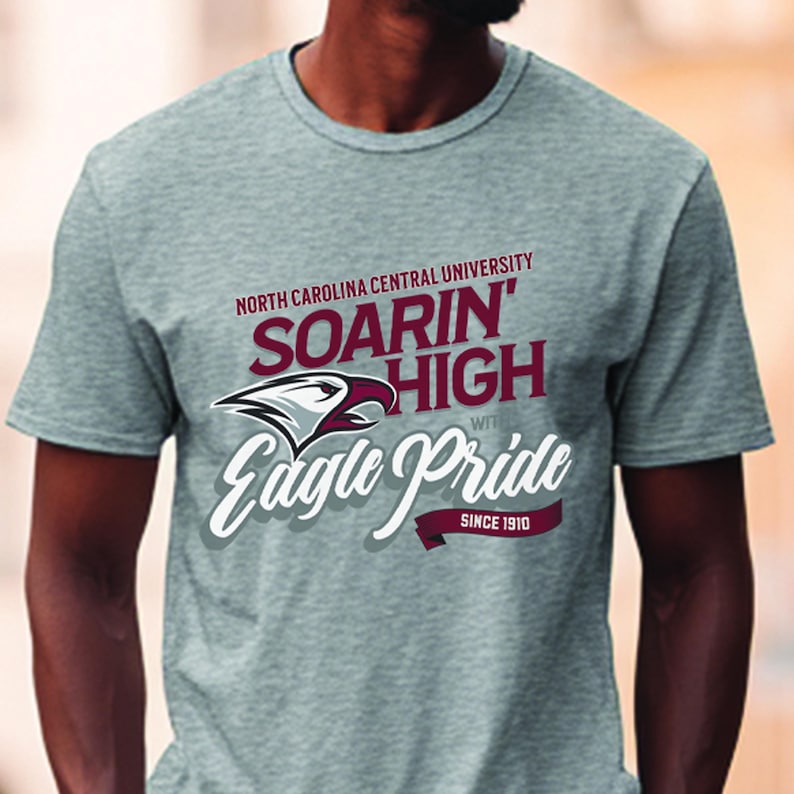 Peut inclure: T-shirt gris avec le texte "NORTH CAROLINA CENTRAL UNIVERSITY" au-dessus de "SOARIN' HIGH" et "Eagle Pride" avec un graphisme d'aigle. Le t-shirt porte &eacute;galement l'inscription "SINCE 1910".