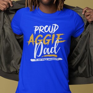 May include: Royal blue t-shirt with the text "PROUD AGGIE Dad" in white and gold lettering. The text "NC A&T STATE UNIVERSITY" is printed in white at the bottom. The shirt is a crew neck style.