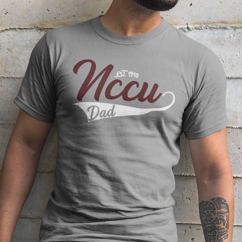 May include: A grey t-shirt with a white baseball-style graphic that says "NCCU Dad" in maroon lettering with "EST. 1910" above the word "NCCU".