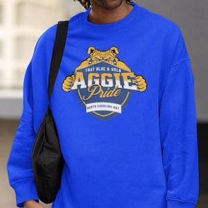 Pode incluir: Camisola azul royal com um gr&aacute;fico que diz "That Blue & Gold Aggie Pride North Carolina A&T". O design apresenta um mascote de bulldog em ouro e azul. A camisola &eacute; de gola redonda.