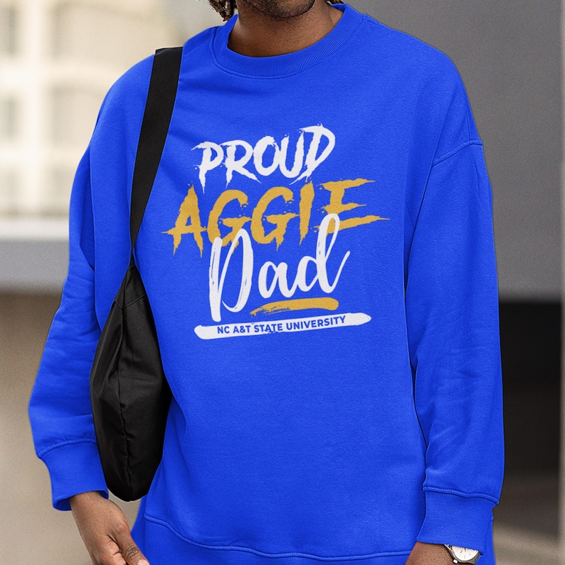 May include: Royal blue sweatshirt with the text "PROUD AGGIE Dad" in white and gold lettering. The text "NC A&T STATE UNIVERSITY" is below the main text. The sweatshirt is a crew neck style.