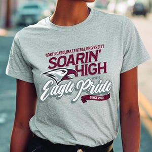 Peut inclure: Un t-shirt gris clair avec le texte "North Carolina Central University" et "Soarin' High with Eagle Pride" en bordeaux et blanc. Le t-shirt pr&eacute;sente &eacute;galement un aigle et le texte "Since 1910".