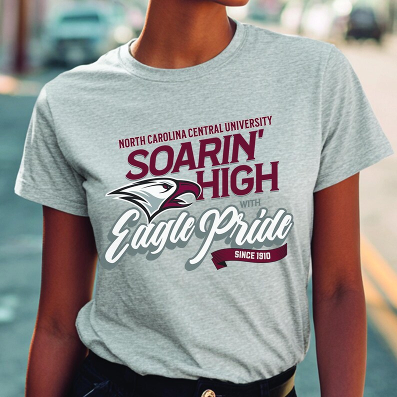 Peut inclure: Un t-shirt gris clair avec le texte "North Carolina Central University" et "Soarin' High with Eagle Pride" en bordeaux et blanc. Le t-shirt pr&eacute;sente &eacute;galement un aigle et le texte "Since 1910".