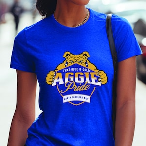 May include: Royal blue t-shirt featuring a gold and blue graphic with the text "THAT BLUE & GOLD AGGIE Pride NORTH CAROLINA A&T". The graphic includes a bulldog mascot. The shirt is short-sleeved.