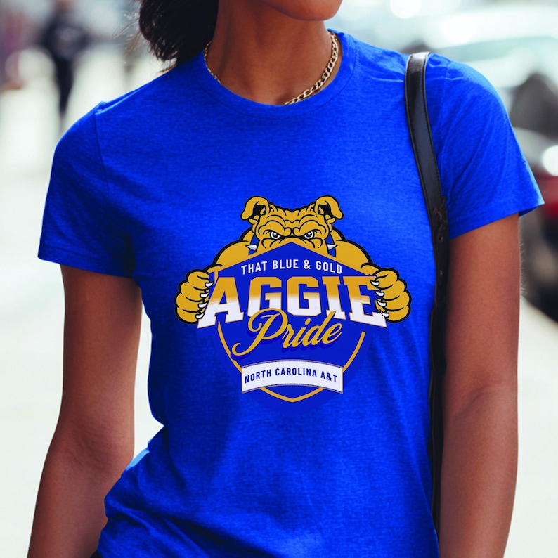 May include: Royal blue t-shirt featuring a gold and blue graphic with the text "THAT BLUE & GOLD AGGIE Pride NORTH CAROLINA A&T". The graphic includes a bulldog mascot. The shirt is short-sleeved.