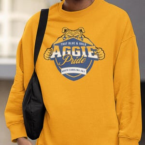 Pode incluir: Camisola mostarda com um gr&aacute;fico que diz "THAT BLUE & GOLD AGGIE Pride NORTH CAROLINA A&T". O gr&aacute;fico apresenta um buldogue e um design de escudo. A camisola &eacute; um estilo de gola redonda.
