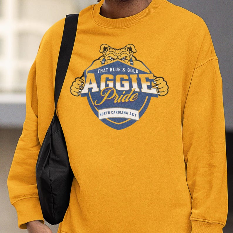 Pode incluir: Camisola mostarda com um gr&aacute;fico que diz "THAT BLUE & GOLD AGGIE Pride NORTH CAROLINA A&T". O gr&aacute;fico apresenta um buldogue e um design de escudo. A camisola &eacute; um estilo de gola redonda.