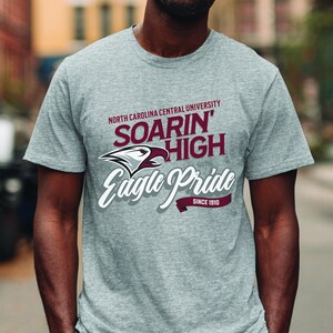 Peut inclure: Un t-shirt gris chin&eacute; avec l'inscription "North Carolina Central University", "Soarin' High with Eagle Pride Since 1910". Le design comprend un aigle blanc et bordeaux.