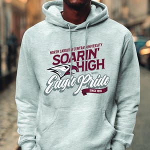 May include: A light gray hoodie with a drawstring hood and a kangaroo pocket. The front features text that reads "North Carolina Central University Soarin' High with Eagle Pride Since 1910" in maroon and white.