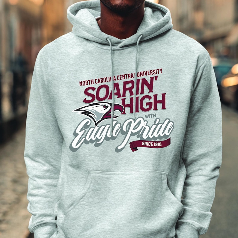 May include: A light gray hoodie with a drawstring hood and a kangaroo pocket. The front features text that reads "North Carolina Central University Soarin' High with Eagle Pride Since 1910" in maroon and white.