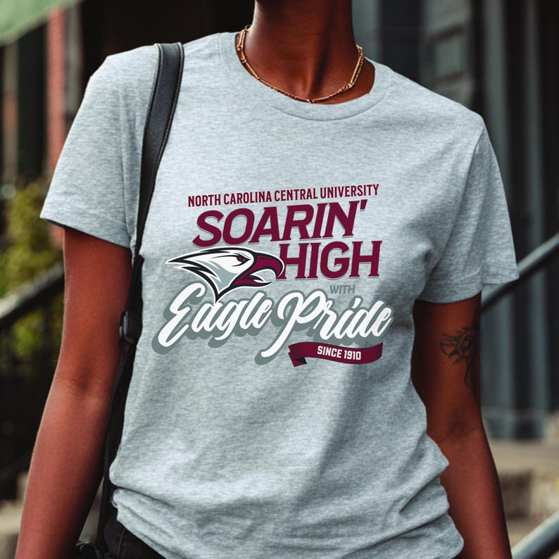 Peut inclure: Un t-shirt gris chin&eacute; avec le texte "NORTH CAROLINA CENTRAL UNIVERSITY" et "SOARIN' HIGH with Eagle Pride SINCE 1910" en bordeaux et blanc. Le t-shirt pr&eacute;sente un motif d'aigle.