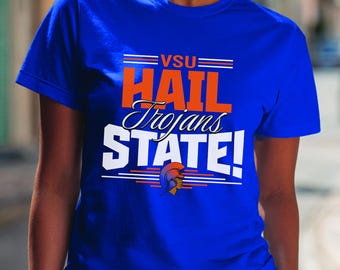 Può includere: T-shirt blu reale con la scritta "VSU HAIL Trojans STATE!" in arancione e bianco. La maglietta presenta una grafica di un elmo troiano. Una maglietta a maniche corte e girocollo.
